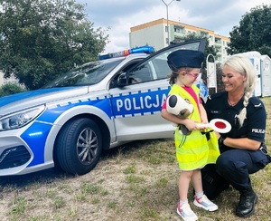 Policjantka pozuje z dzieckiem na tle radiowozu. Dziecko ma na sobie kamizelkę odblaskową i czapkę policyjną.