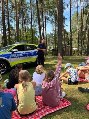 Policjant z grupą dzieci podczas prelekcji w tle radiowóz.