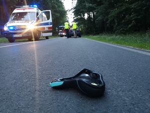 Miejsce wypadku. Na pierwszym planie na drodze leży siodełko od roweru, w tle karetka pogotowia i wóz strażacki na drodze oraz dwaj policjanci przy samochodzie osobowym