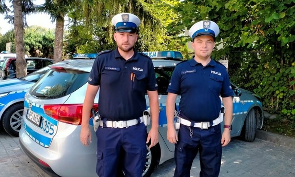 na zdjęciu dwóch umundurowanych funkcjonariuszy stoi przy radiowozie