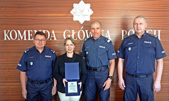 Zdjęcie pamiątkowe nadinsp. Romana Kustera oraz Pani Konsul Hejduk-Gromek trzymającej w ręku okolicznościowy ryngraf na tle godła i napisu Komenda Główna Policji w towarzystwie Dyrektora Biura Prewencji KGP insp. Roberta Kumora