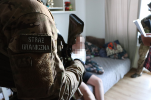 na zdjęciu funkcjonariusze straży granicznej stoją w pomieszczeniu, na łóżku siedzi zatrzymany mężczyzna