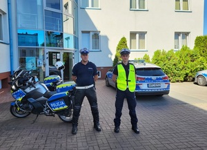 dwaj policjanci stoją przed budynkiem komendy Policji, za nimi stoją dwa motocykle i radiowóz