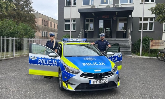 Na zdjęciu widoczny radiowóz policyjny zaparkowany  przed budynkiem, obok pojazdu stoją funkcjonariusze policji
