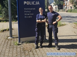 dwoje umundurowanych policjantów stojących przy tablicy informacyjnej z napisem Komisariat Policji Wrocław Osiedle
