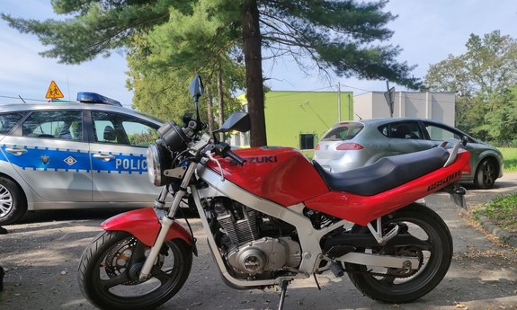 na zdjęciu motocykl oraz radiowóz na poboczu w tle budynek