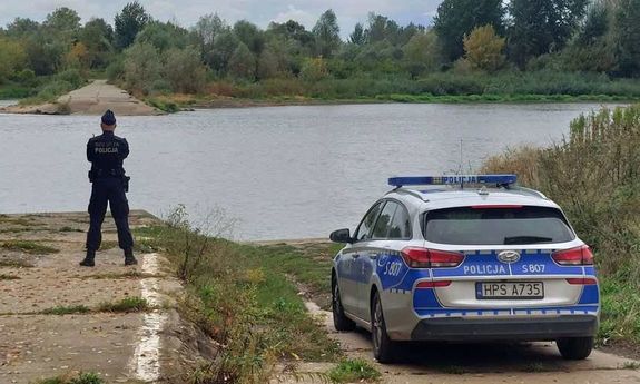 policjant stoi przy radiowozie. przed nim płynie rzeka