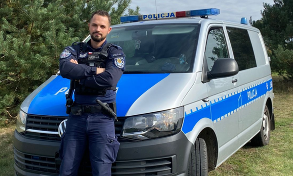 Na zdjęciu umundurowany funkcjonariusz stoi na przeciwko radiowozu.
