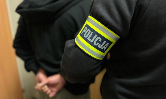 Funkcjonariusz z opaską z napisem policja prowadzi zatrzymanego.