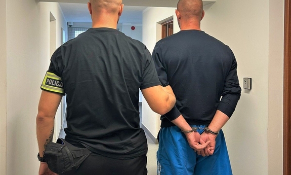 Na zdjęciu policjant pionu kryminalnego trzyma prowadzi, który ma założone kajdanki na ręce.