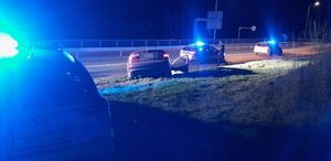 Na zdjęciu dwa oznakowane radiowozy, samochód służbowy na ulicy i pojazd, którym kierowca uciekał przed kontrolą drogową. Obok samochodu stoi policjant.