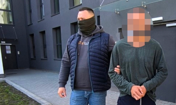 Funkcjonariusz prowadzi zatrzymanego mężczyznę zakutego w kajdanki zespolone.