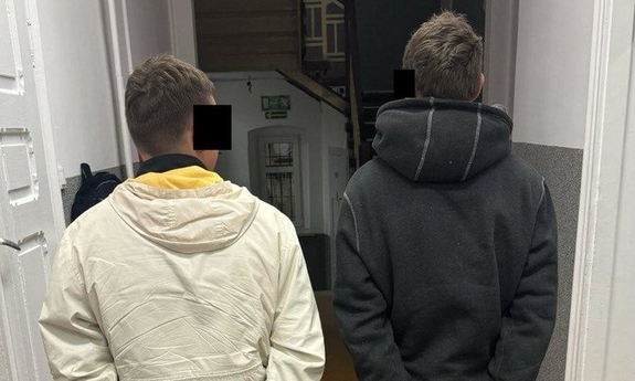 Na zdjęciu dwóch zatrzymanych mężczyzn stoi na korytarzu z kajdankami na rękach.