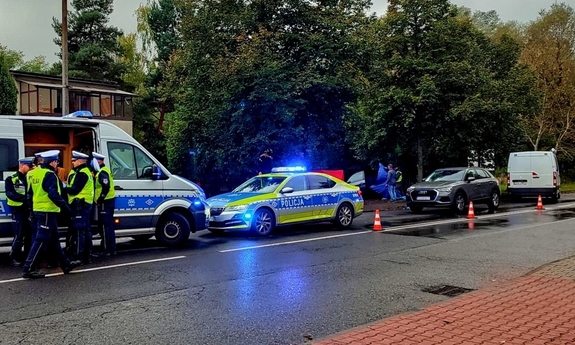 Na zdjęciu ulica na której stoją radiowozy, widać grupę policjantów przy furgonie policyjnym w tle na poboczu rozbite auto a przy nim ludzie i parawan.