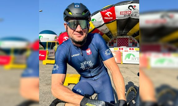 Policjant na rowerze w kolarskim stroju pozuje do zdjęcia, z tyłu widoczne są plakaty sponsorów wyścigu i organizatora.