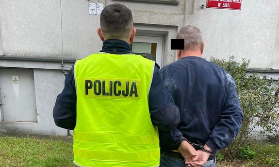 Mężczyzna z założonymi kajdankami na ręce trzymane z tyłu jest prowadzony przez umundurowanego policjanta do komisariatu.