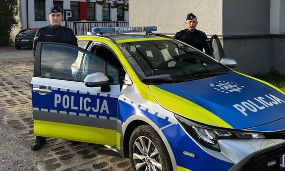 Na zdjęciu dwóch umundurowanych funkcjonariuszy stoi przy oznakowanym radiowozie.