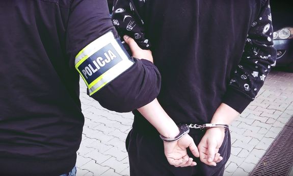 Nieumundurowany policjant, w opasce na ręce z napisem Policja, prowadzi zatrzymanego. Widok z tyłu.