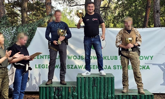 Na zdjęciu uczestnicy zawodów strzeleckich stoi na podium po zakończonym turnieju. Mężczyźni trzymają w ręku puchar.