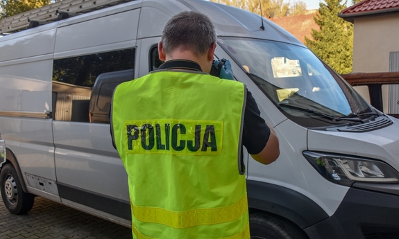 Na zdjęciu policjant w odblaskowej kamizelce z napisem Policja fotografuje odzyskanego białego busa, który stoi na posesji.