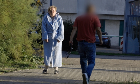 Starsza kobieta w szlafroku na ulicy. Obok idzie mężczyzna w przeciwnym kierunku.