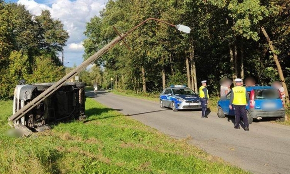 Miejsce zdarzenia. W rowie leży przewrócony samochód, obok zaparkowany radiowóz policyjny i samochód osobowy, przy którym stoją policjanci i mężczyźni.