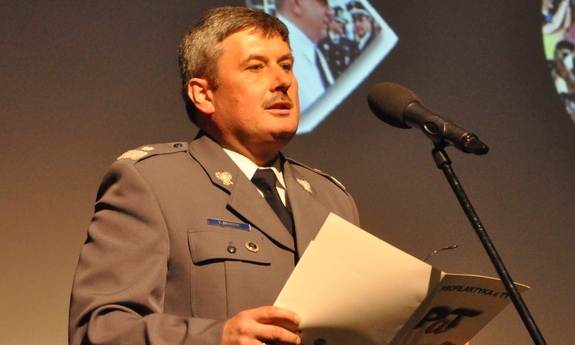 Gen. Tadeusz Budzik stoi na scenie przy mikrofonie przemawiając do publiczności. w dłoniach trzyma otwartą teczkę z napisem Profilaktyka a Ty i logiem PaT.
