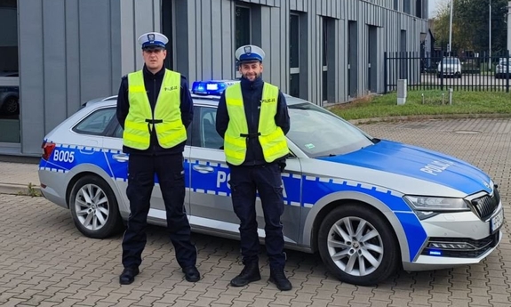 Dwaj policjanci w kamizelkach odblaskowych stojący koło radiowozu przed budynkiem policji.