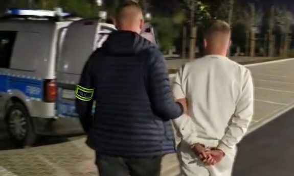 Nieumundurowany policjant prowadzi zatrzymanego mężczyznę w kierunku oznakowanego radiowozu.
