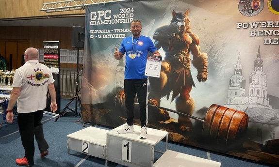 Na zdjęciu mężczyzna stoi na podium z medalem na szyi i dyplomem w ręce.