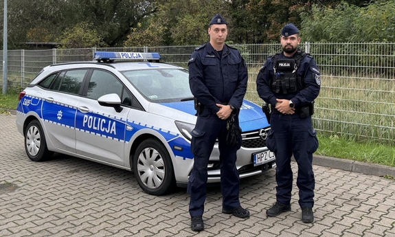 Na zdjęciu dwóch umundurowanych policjantów stoi przy radiowozie.