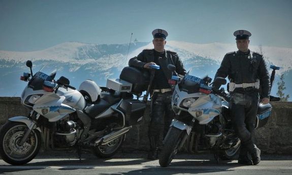 Dwaj umundurowani policjanci drogówki stoją przy motocyklach. W tle widać góry.