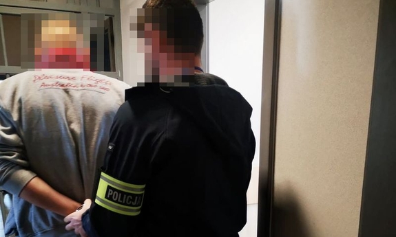 Na zdjęciu policjant z odblaskową naszywką na przedramieniu prowadzi zatrzymanego mężczyznę.