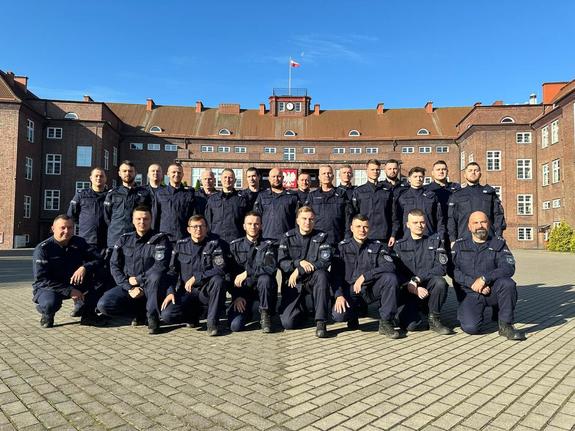 Funkcjonariusze Samodzielnego Pododdziału Prewencji Policji (SPPP) w Legnicy w mundurach, w dwuszeregu pozują do zdjęcia. Pierwszy rząd kuca, drugi stoi. W tle widoczny jest budynek.