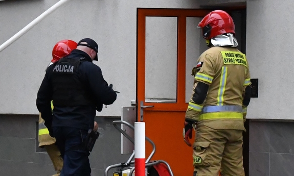 Policjant i dwaj strażacy przed budynkiem gdzie w jednym z mieszkań wybuchł pożar.