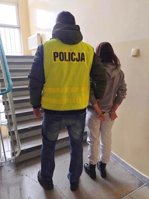 Na zdjęciu policjant w odblaskowej kamizelce prowadzi zatrzymaną kobietę po klatce schodowej. Kobieta ma na nadgarstkach kajdanki.