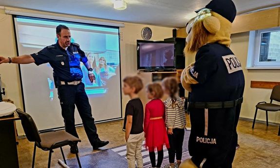 policjant pokazuje coś na tablicy trójce dzieci w towarzystwie policyjnej maskotki.