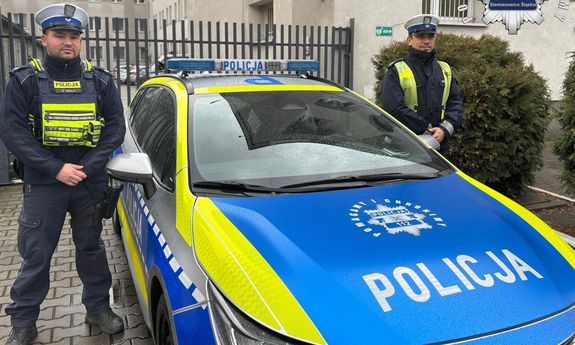 dwaj umundurowani Policjanci stoją obok radiowozu.