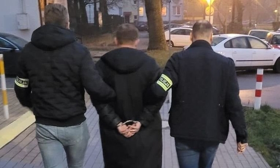 Na zdjęciu dwóch policjantów prowadzi zatrzymanego mężczyznę w kajdankach.