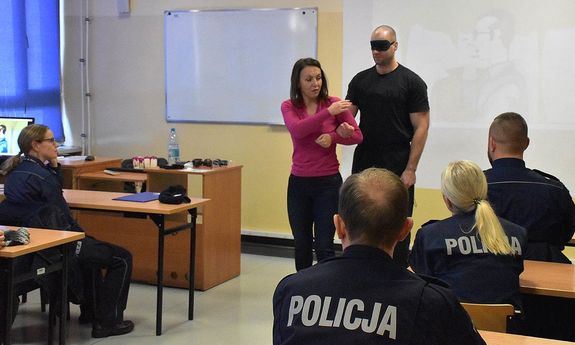 policjanci siedzą na sali, przed nimi stoi kobieta, która prowadzi policjanta z opaska na oczach.