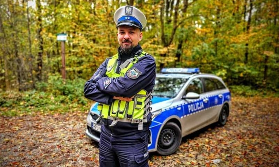 Na zdjęciu umundurowany policjant, w tle oznakowany radiowóz policyjny.