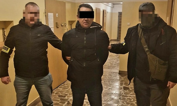 Na zdjęciu dwóch policjantów prowadzi zatrzymanego mężczyznę w kajdankach.