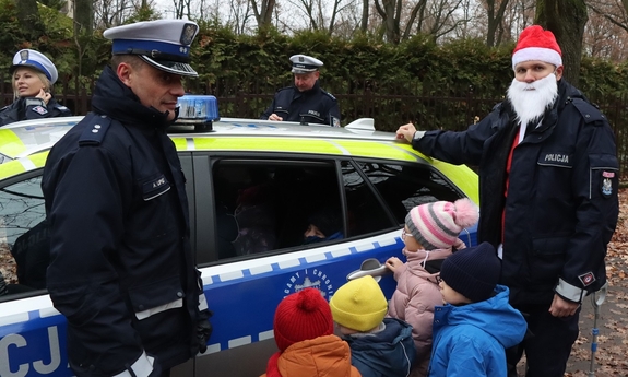Czworo dzieci przy radiowozie policyjnym razem z policjantami - jeden z nich ma na głowie czapkę Mikołaja.