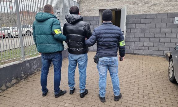 dwóch policjantów ubranych po cywilnemu prowadzi mężczyznę, który ma założone kajdanki na ręce trzymane z przodu.