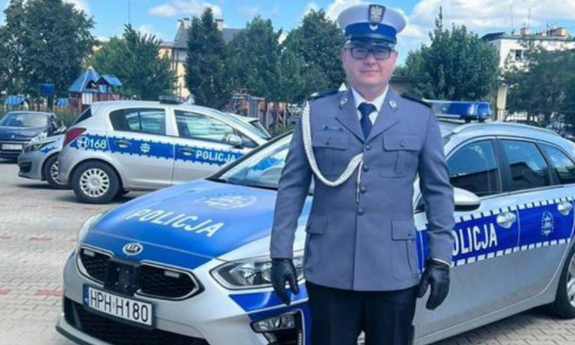 Na zdjęciu umundurowany policjant. W tle stoją zaparkowane oznakowane radiowozy.