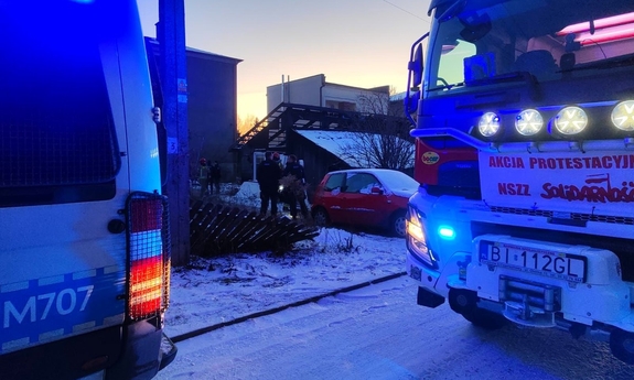 Fragment wozu strażackiego i radiowozu policyjnego. W głębi zabudowania i stojący policjanci ze strażakami.