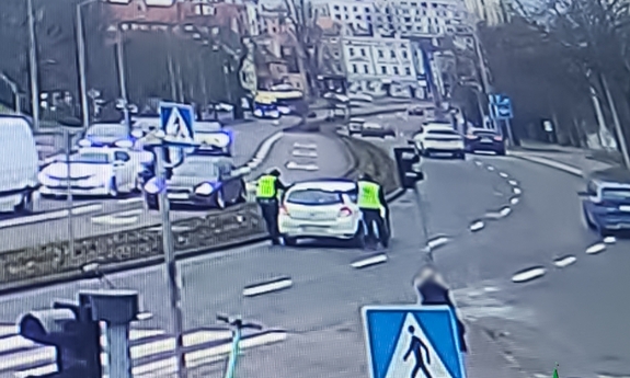 Widok na ulicę. Umundurowani policjanci pomagają kobiecie w białym samochodzie.