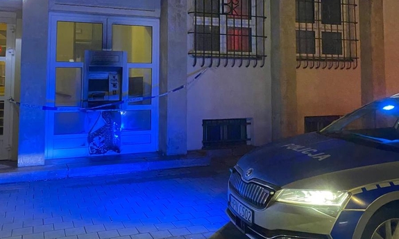 Uszkodzony bankomat w ścianie budynku. Obok radiowóz.