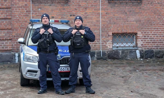 Dwóch umundurowanych policjantów stoi przy oznakowanym radiowozie.