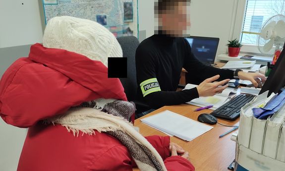 Siedzący za biurkiem ubrany po cywilnemu policjant a przy nim w czapce i kurtce zatrzymana kobieta.